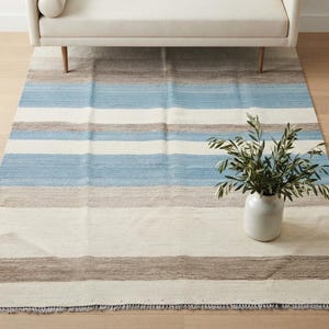 Op de afbeelding: Een rechthoekig vloerkleed met horizontale strepen in tinten lichtblauw, beige en gebroken wit. Het kleed heeft een geweven textuur met een franjerand aan de onderkant en een klein, subtiel stervormig detail in de rechterbenedenhoek.