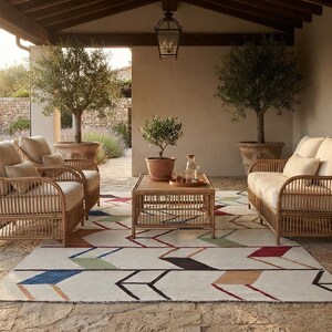 May include: A patio furniture set consisting of two armchairs and a sofa with light beige cushions, arranged around a rectangular wooden coffee table. The furniture sits on a large area rug featuring a repeating geometric arrow pattern in blue, green, red, and tan.