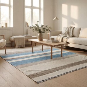 Op de afbeelding: Een rechthoekig vloerkleed met horizontale strepen in tinten lichtblauw, beige, cr&egrave;me en donkerbruin. Het kleed heeft een platgeweven textuur met korte franjes langs de randen en ligt op een lichte houten vloer.