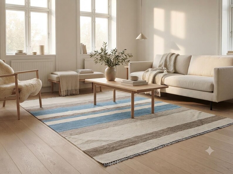 Op de afbeelding: Een rechthoekig vloerkleed met horizontale strepen in tinten lichtblauw, beige, cr&egrave;me en donkerbruin. Het kleed heeft een platgeweven textuur met korte franjes langs de randen en ligt op een lichte houten vloer.