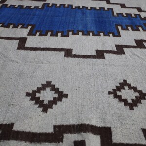 May include: A white and brown patterned rug with a blue stripe across the top. The rug has a geometric design with diamond shapes and a border.