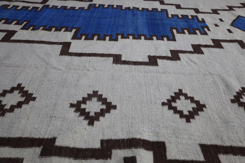 May include: A white and brown patterned rug with a blue stripe across the top. The rug has a geometric design with diamond shapes and a border.