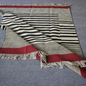 May include: A black and white striped woven rug with a red border and fringe. The rug is folded in half and the fringe is visible on both sides.