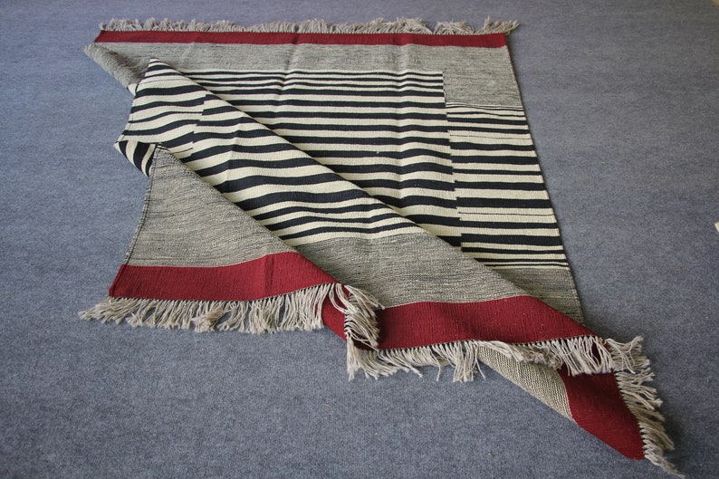 May include: A black and white striped woven rug with a red border and fringe. The rug is folded in half and the fringe is visible on both sides.
