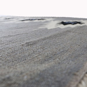 May include: A close-up of a grey woven rug with a fringed border. The rug has a subtle pattern of white and black geometric shapes.