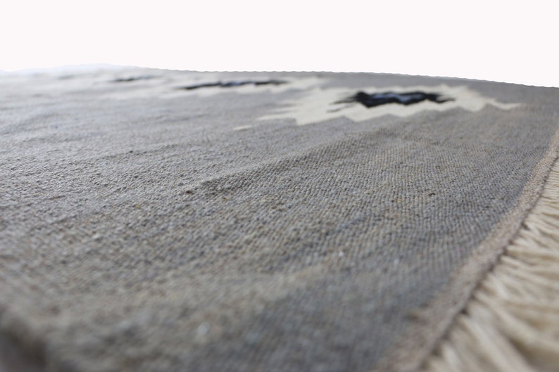 May include: A close-up of a grey woven rug with a fringed border. The rug has a subtle pattern of white and black geometric shapes.