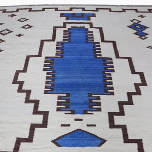 May include: A white rug with a geometric pattern in brown and blue. The rug has a central design of a large blue rectangle with brown borders.