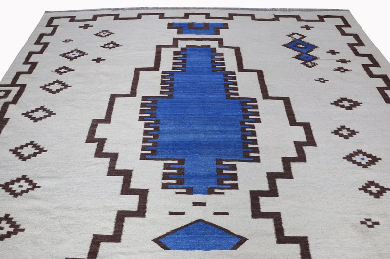 May include: A white rug with a geometric pattern in brown and blue. The rug has a central design of a large blue rectangle with brown borders.