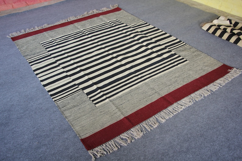 May include: A black and white striped rug with a red border and fringe. The rug is made of woven fabric and has a geometric pattern.