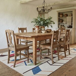 May include: A rectangular wooden dining table with four matching chairs featuring carved backrests. The table is set on a large area rug with a white base and a geometric pattern of blue, red, and green lines. A ceramic pitcher and bowl sit on the table.