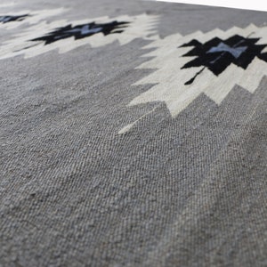 May include: A grey woven rug with a white and black geometric pattern. The rug has a textured surface.