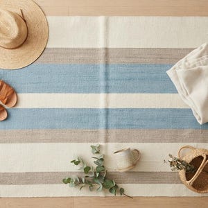 Op de afbeelding: Een rechthoekig geweven kleed met horizontale strepen in cr&egrave;me, lichtblauw en beige met franjes aan de randen. Het kleed is omringd door een strooien hoed, een paar bruine leren sandalen, een gevouwen cr&egrave;mekleurige stof, een mok en een klein mandje.