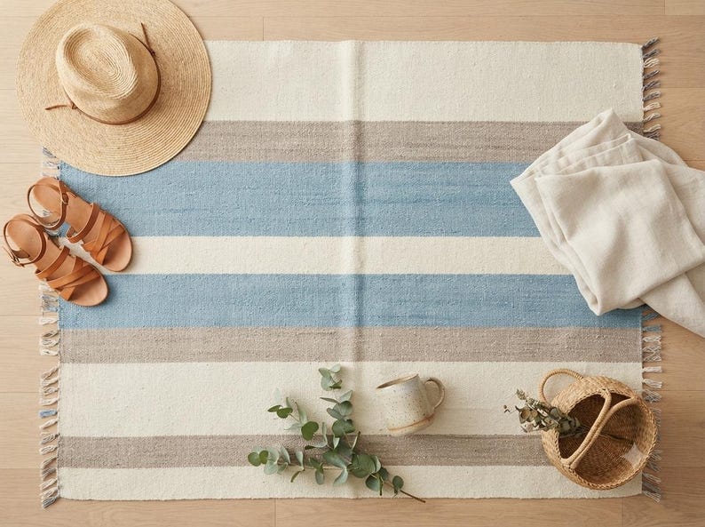 Op de afbeelding: Een rechthoekig geweven kleed met horizontale strepen in cr&egrave;me, lichtblauw en beige met franjes aan de randen. Het kleed is omringd door een strooien hoed, een paar bruine leren sandalen, een gevouwen cr&egrave;mekleurige stof, een mok en een klein mandje.