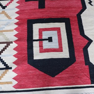Puede incluir: Primer plano de una alfombra tejida con un patr&oacute;n geom&eacute;trico en tonos rojos, negros, blancos y marrones. La alfombra tiene un borde con flecos.