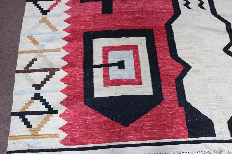 Puede incluir: Primer plano de una alfombra tejida con un patr&oacute;n geom&eacute;trico en tonos rojos, negros, blancos y marrones. La alfombra tiene un borde con flecos.