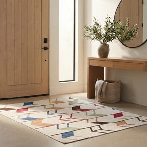 May include: A rectangular area rug featuring a cream-colored background with a repeating geometric pattern of interconnected lines and shapes in shades of red, blue, brown, and light green, arranged in a structured, linear, and rhythmic design.