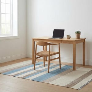Op de afbeelding: Een bureau van licht hout met een lade, een houten stoel met een gevlochten zitting en een laptop erop. Een kleine vetplant in een pot en een keramische mok staan op het bureau. Het meubilair staat op een rechthoekig vloerkleed met blauwe strepen.