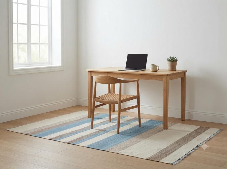 Op de afbeelding: Een bureau van licht hout met een lade, een houten stoel met een gevlochten zitting en een laptop erop. Een kleine vetplant in een pot en een keramische mok staan op het bureau. Het meubilair staat op een rechthoekig vloerkleed met blauwe strepen.
