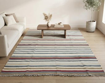 Puede incluir: Una alfombra rectangular con un patrón de rayas horizontales en tonos rojo apagado, azul, beige y crema. La alfombra tiene una textura de tejido plano e incluye detalles de flecos cortos y deshilachados en los dos bordes más cortos.
