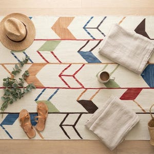 May include: A rectangular area rug featuring a repeating arrow pattern in shades of blue, green, red, brown, and tan on an off-white background. The rug includes fringe on the short ends and is decorated with a straw hat, sandals, two folded cloths, a mug, a sprig of eucalyptus, and a woven basket.