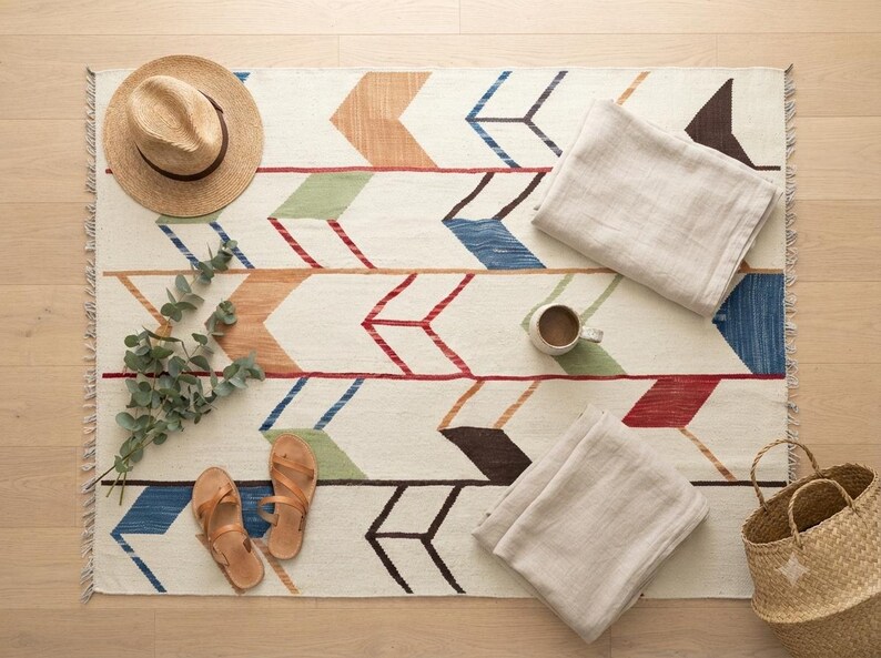 May include: A rectangular area rug featuring a repeating arrow pattern in shades of blue, green, red, brown, and tan on an off-white background. The rug includes fringe on the short ends and is decorated with a straw hat, sandals, two folded cloths, a mug, a sprig of eucalyptus, and a woven basket.