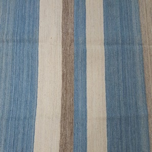 Op de afbeelding: Een geweven textielkleed met een herhalend verticaal streeppatroon in tinten lichtblauw, gebroken wit en een gedempt aards bruin. De stof vertoont een zichtbaar, getextureerd weefsel met consistente evenwijdige lijnen over het oppervlak.