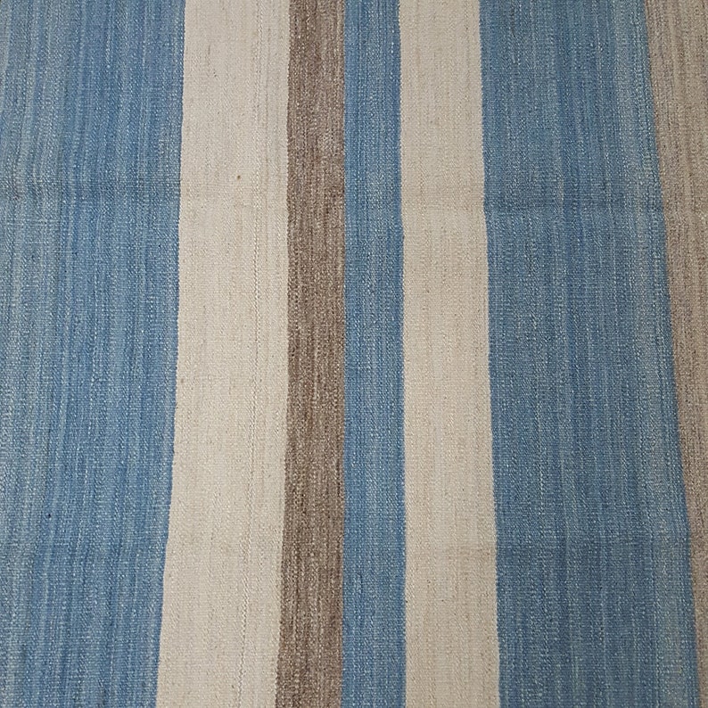 Op de afbeelding: Een geweven textielkleed met een herhalend verticaal streeppatroon in tinten lichtblauw, gebroken wit en een gedempt aards bruin. De stof vertoont een zichtbaar, getextureerd weefsel met consistente evenwijdige lijnen over het oppervlak.