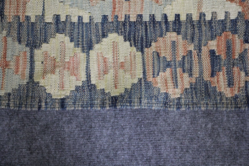 May include: Close-up of a woven rug with a blue, beige and orange geometric pattern. The rug has a fringe border and is made of a soft, textured material.