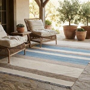 Op de afbeelding: Een rechthoekig vloerkleed met horizontale strepen in tinten lichtblauw, beige en donkerbruin. Het kleed heeft een geweven textuur met korte franjes langs de randen, geplaatst op een stenen terras onder twee rotan stoelen met kussens.