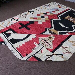 Puede incluir: Una alfombra de color blanco, rojo, negro y marr&oacute;n con dise&ntilde;os geom&eacute;tricos. La alfombra est&aacute; doblada por la mitad y tiene un borde con flecos.