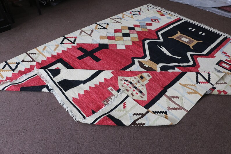 Puede incluir: Una alfombra de color blanco, rojo, negro y marr&oacute;n con dise&ntilde;os geom&eacute;tricos. La alfombra est&aacute; doblada por la mitad y tiene un borde con flecos.