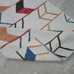 May include: A woven area rug featuring an off-white base with a repeating geometric arrow pattern in shades of dark brown, red, orange, blue, and light green. The rug is partially folded, revealing the textured weave and the colorful design.