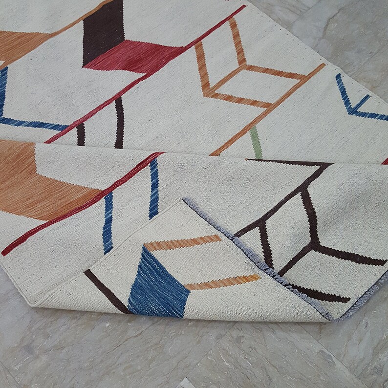 May include: A woven area rug featuring an off-white base with a repeating geometric arrow pattern in shades of dark brown, red, orange, blue, and light green. The rug is partially folded, revealing the textured weave and the colorful design.