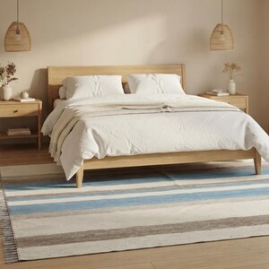 Op de afbeelding: Een rechthoekig vloerkleed met horizontale strepen in de kleuren lichtblauw, beige, grijs en wit. Het kleed heeft een geweven textuur met korte franjes langs de randen en ligt op een lichte houten vloer in een slaapkamer.