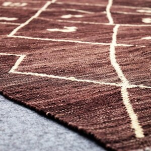 May include: A close-up of a brown woven rug with a white geometric pattern. The rug has a fringe on the bottom edge.