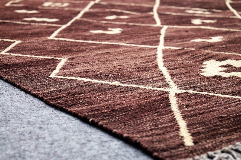 May include: A close-up of a brown woven rug with a white geometric pattern. The rug has a fringe on the bottom edge.