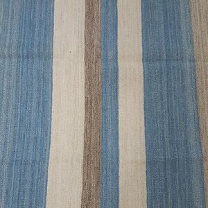 Op de afbeelding: Een geweven textielkleed met een herhalend verticaal streeppatroon in tinten lichtblauw, gebroken wit en een gedempt aards bruin. De stof vertoont een zichtbaar, getextureerd weefsel met consistente evenwijdige lijnen over het oppervlak.