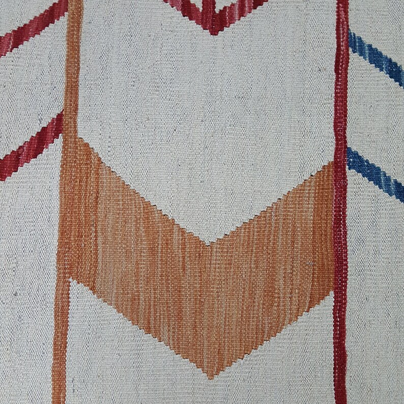 May include: A woven textile featuring a cream-colored base with a prominent orange chevron pattern in the center. The design includes vertical red and blue lines that frame the central shape, creating a geometric composition with visible fabric texture.