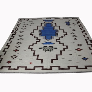 May include: A white rug with a geometric pattern in shades of blue, red, and brown. The rug has a central design with a blue diamond shape and a black and white geometric pattern.