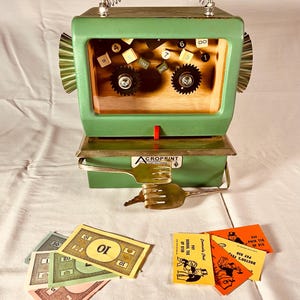 May include: A green, retro-style art piece resembling a calculator or cash register. It features a spring handle, gears, and a wooden interior with number keys. Prop money and game cards are arranged in front.
