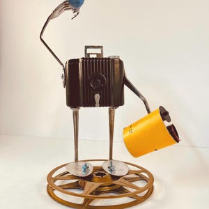 May include: A black vintage camera with a yellow film canister and a blue glass ball on top, standing on a gold film reel.