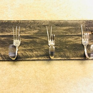 May include: Three silver forks are mounted on a dark brown wooden board to create a rustic coat rack.