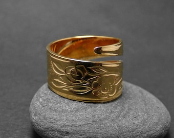 May include: A gold-colored ring with a floral design, resting on a gray stone against a dark background. The ring features an engraved pattern of flowers and leaves. The ring's design is open, with a gap on one side.