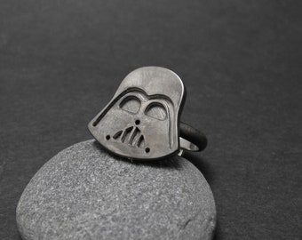 May include: A silver ring featuring a Darth Vader helmet design. The ring is resting on a grey stone.
