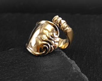 May include: A gold-toned ring with a unique design. The ring is made of a single piece of metal that is bent into a curved shape. The ring is set on a black background.