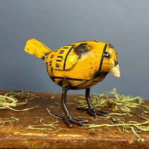 May include: A handcrafted metal bird sculpture with a yellow body and black stripes. The bird has metal legs and a white beak. The body has the word "Infla" printed on it. The sculpture is on a wooden surface.