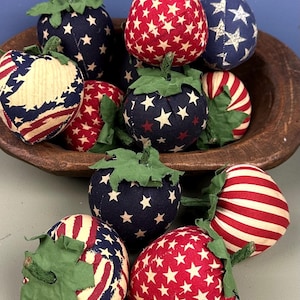 May include: A collection of patriotic fabric strawberries in a wooden bowl. The strawberries feature red, white, and blue patterns, including stars and stripes. Each strawberry has a green fabric stem and leaves.