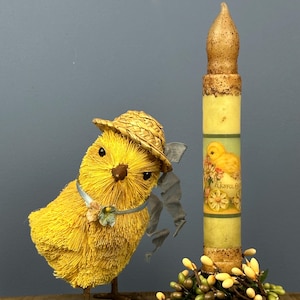 May include: A yellow chick figurine wearing a straw hat and a candle with a chick graphic. The candle has a floral base and a textured top. The chick figurine has a blue ribbon and is on a wooden surface.