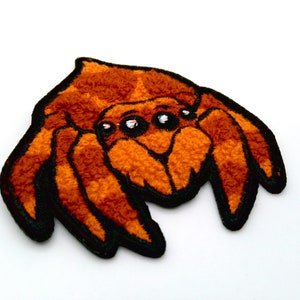 May include: An orange and black embroidered spider patch. The patch features a detailed design of a spider with a fuzzy texture and black outlines. The spider has multiple eyes and is set against a white background.