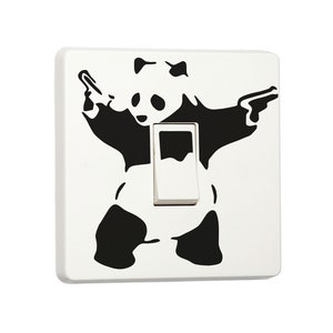 Op de afbeelding: Een witte lichtschakelaar met een sticker van een zwart-witte pandabeer die twee pistolen vasthoudt. De pandabeer draagt ​​een strikje.